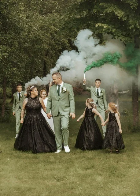 Bruidspaar met hun kinderen en getuigen tussen groene rookfakkels, de bruid in een zwarte jurk